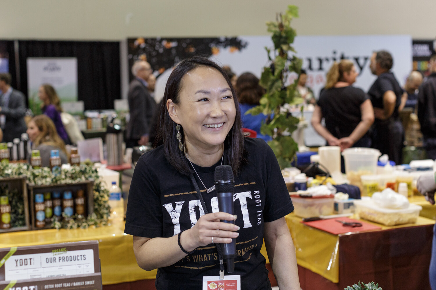 A smiling woman in a black Yoot Root Teas shirt holding a microphone at a booth table.