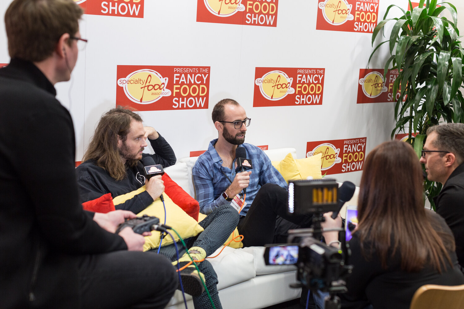 Two men on a white sofa with red and yellow cushions being interviewed.