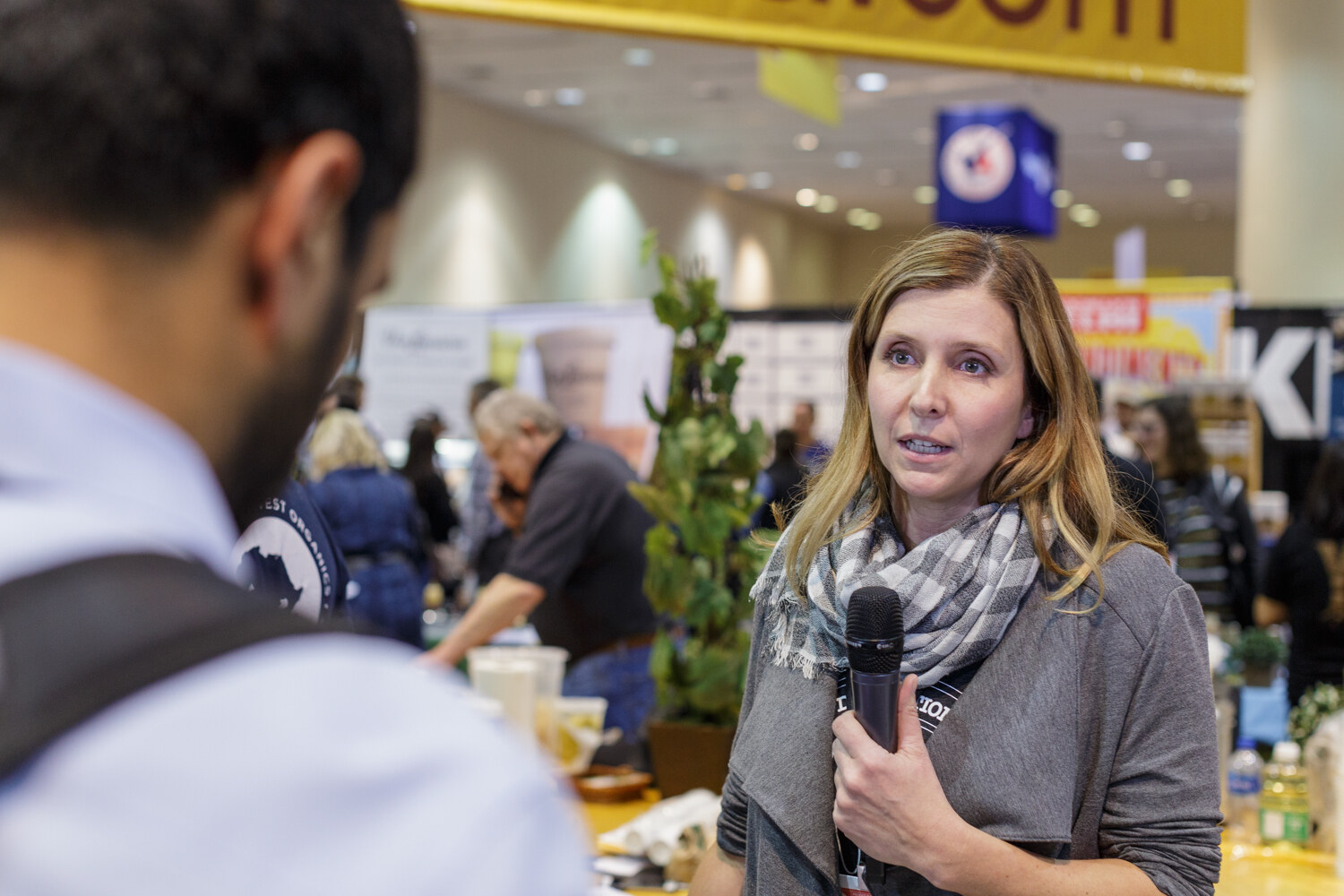 A woman holds a microphone with exhibit booths behind her.