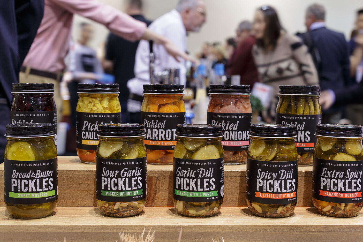 Jars labeled bread and butter, sour garlic, garlic dill and spicy dill pickles with people behind.