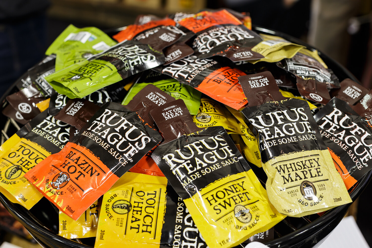 A bowl of sauce packets labeled Rufus Teague in Honey Sweet, Whiskey Maple and other flavors.
