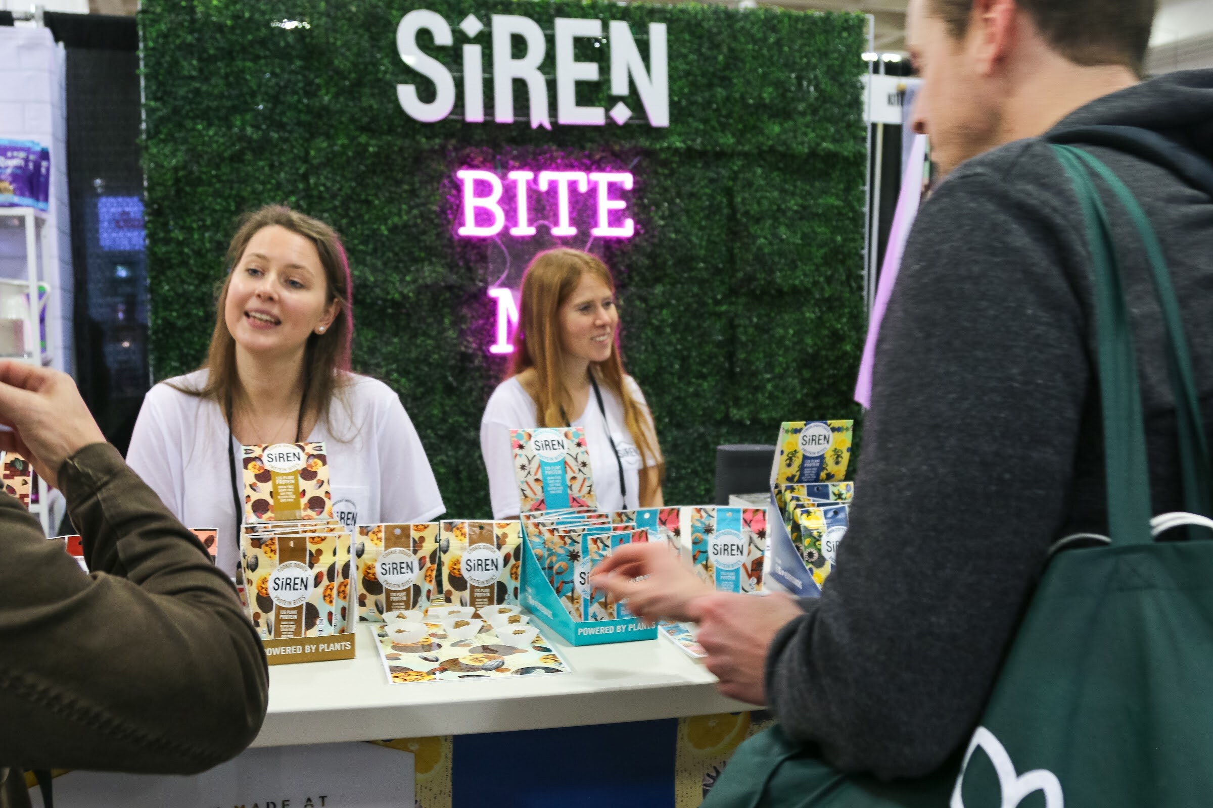 Two women at a booth with a Siren sign and a neon BITE sign, speaking with visitors in the foreground.