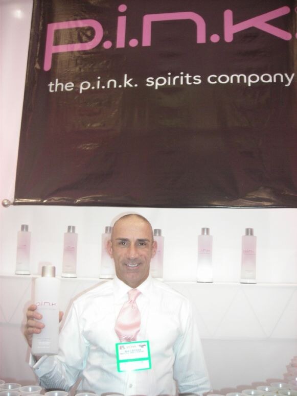A smiling man in a white shirt and pink tie holds a bottle under a p.i.n.k. spirits company banner, with more bottles lined on shelves behind him.