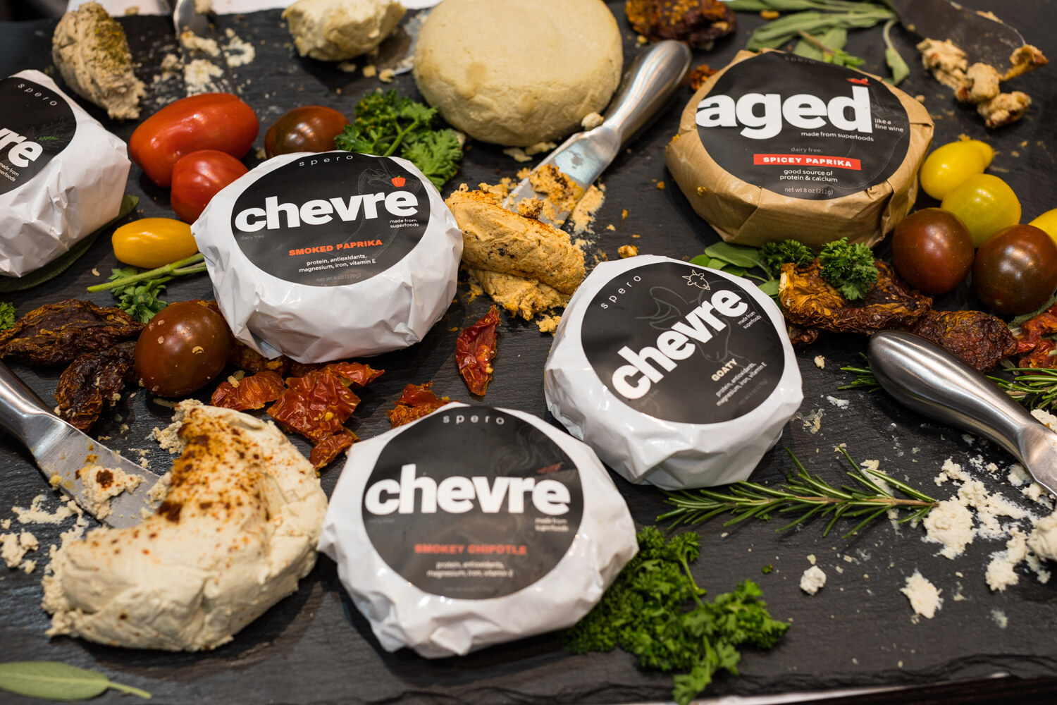 Photo of round wrapped chevre cheeses on a slate board with tomatoes and herbs.