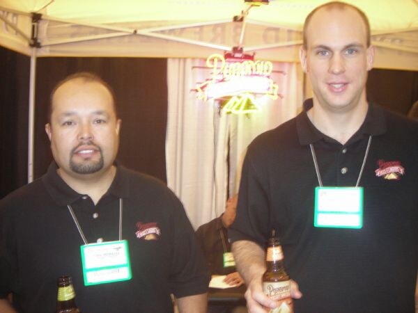 Two men in black Pyramid polo shirts and badges stand under a tent with a neon sign, one holding a Pyramid beer bottle.