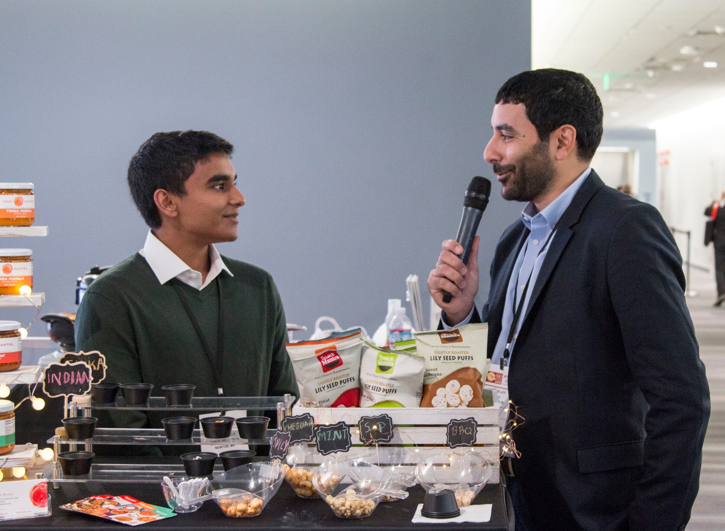 Photo of two men during an interview, one holding a microphone, at a booth of jarred sauces.