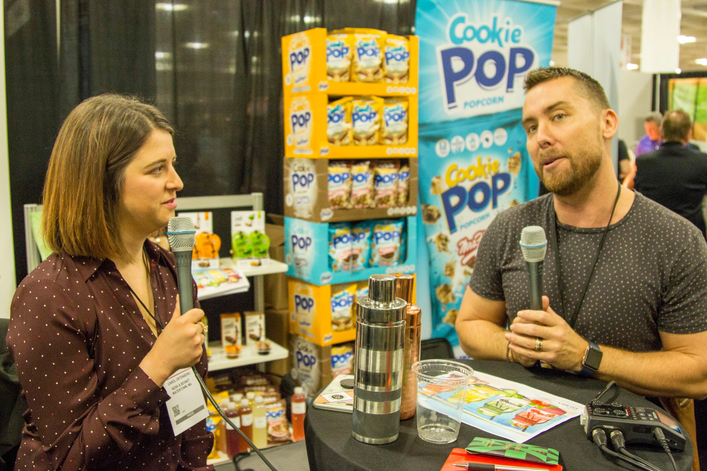 Photo of a woman and a man holding microphones during an interview in front of a Cookie Pop popcorn display.
