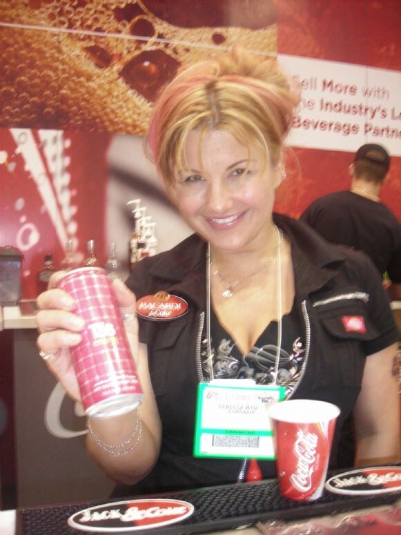 Rebecca Ryu, wearing an exhibitor badge, smiles and holds a pink Tab Energy can behind a bar with a Coca-Cola cup and Jack & Coke bar mat. A Bacardi & Coke pin is on her black top.