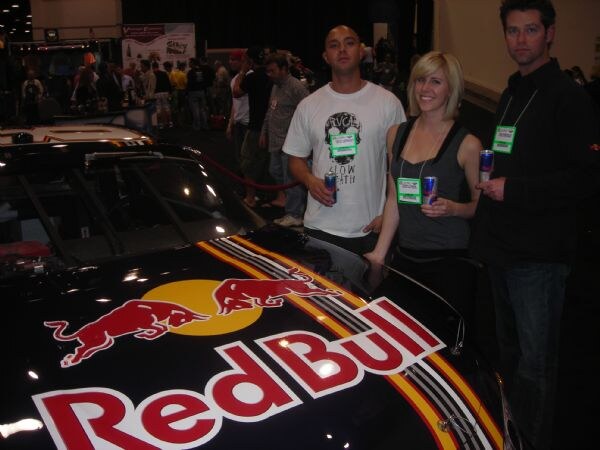 Two men and a woman wearing show badges stand beside a black car with a Red Bull logo on the hood, each holding a Red Bull can. Other attendees are in the background.