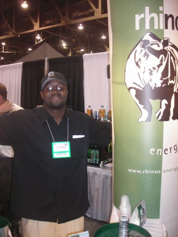 A man in glasses, a cap and a black shirt with a show badge stands with arms outstretched beside a green and white Rhino's Energy banner with a rhinoceros graphic.