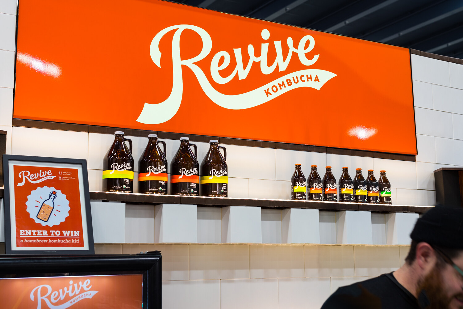 Photo of a bright orange Revive Kombucha sign above a shelf of growler-style bottles.