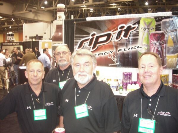 Four men in black polo shirts with Under Armour and Rip It logos and show badges stand together in front of a Rip It Energy Fuel banner showing cans.