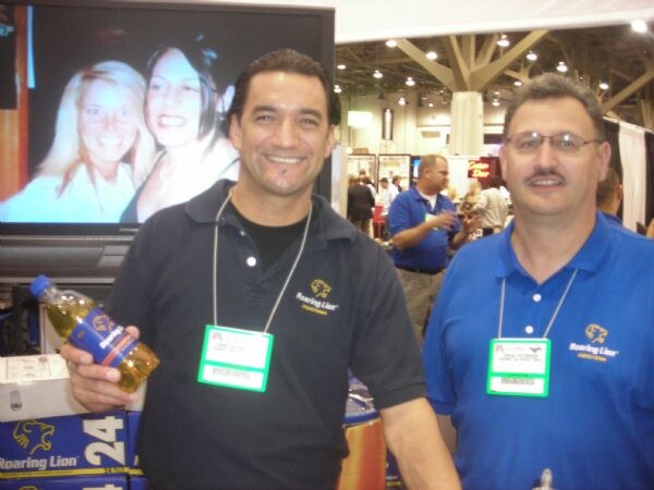 Two smiling men wearing Roaring Lion shirts and show badges stand together; one holds a bottle of drink. A screen and Roaring Lion cases are behind them.