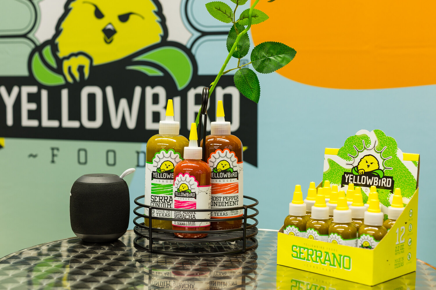 Photo of Yellowbird hot sauce bottles in front of a colorful Yellowbird Foods sign.