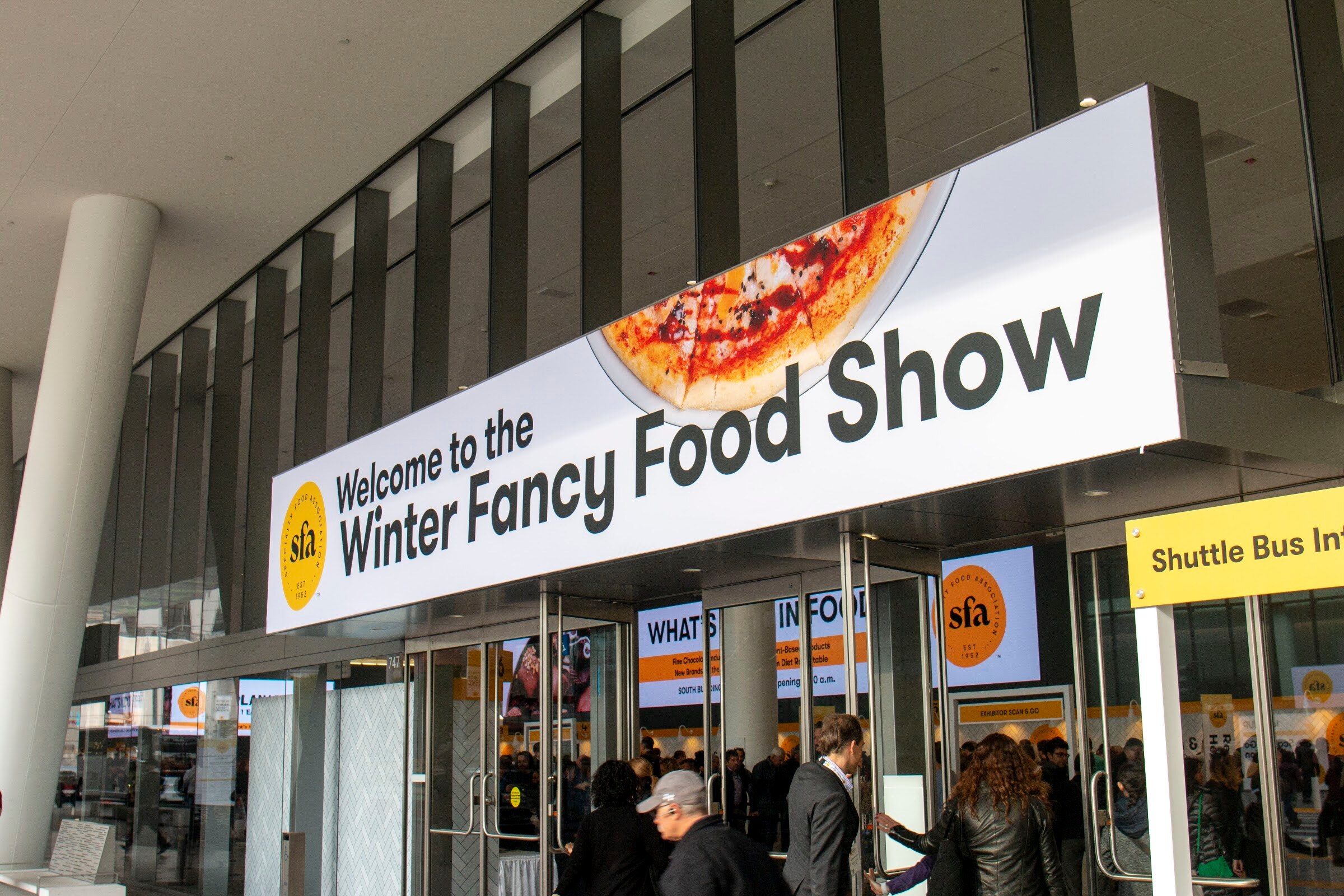 A banner reading Welcome to the Winter Fancy Food Show above glass entrance doors with attendees walking in