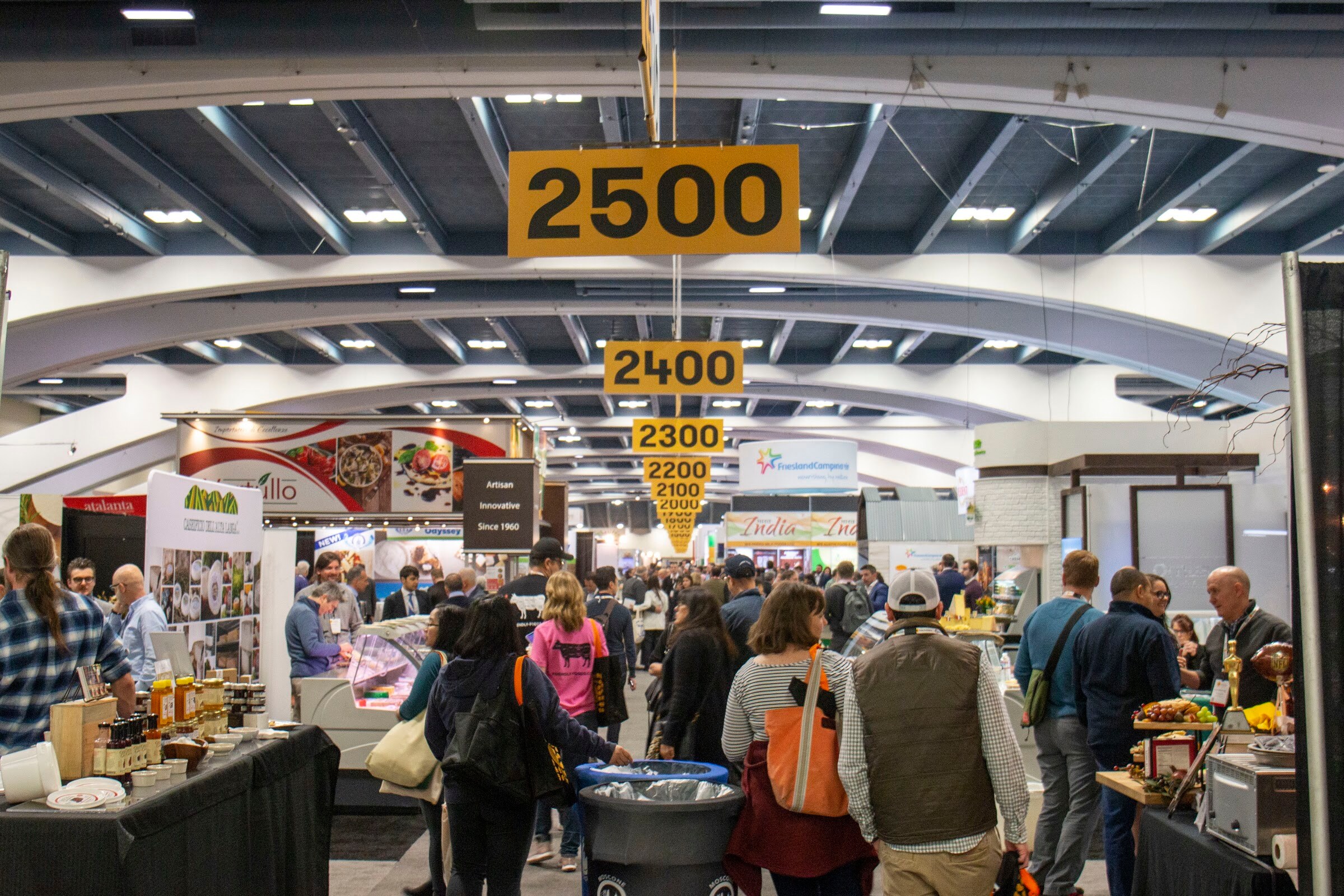A busy exhibit hall with hanging aisle numbers 2300 to 2500 and crowds walking between booths