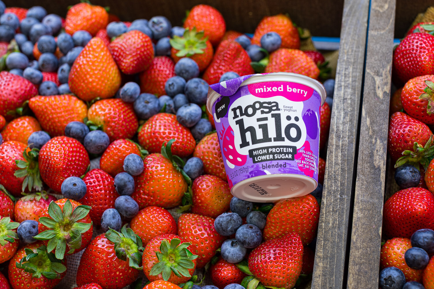 A purple noosa hilö mixed berry yogurt cup resting among fresh strawberries and blueberries