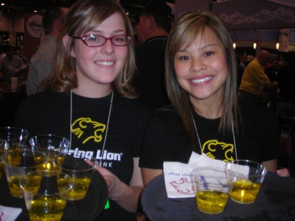 Two smiling women in black Roaring Lion shirts hold trays of small plastic cups filled with a yellow drink.