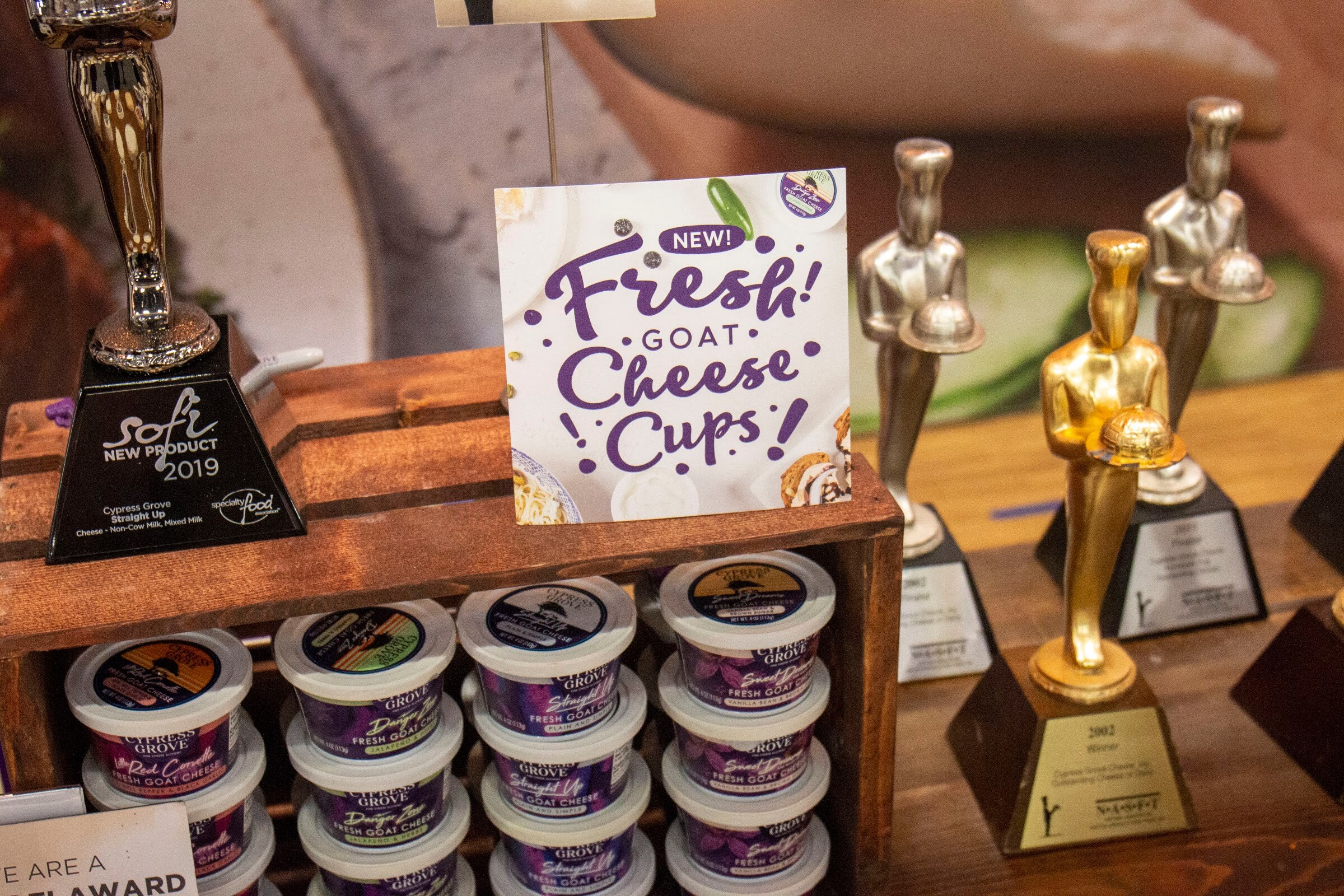 A wooden crate of goat cheese cups next to gold and silver trophies and a sign for new fresh goat cheese cups