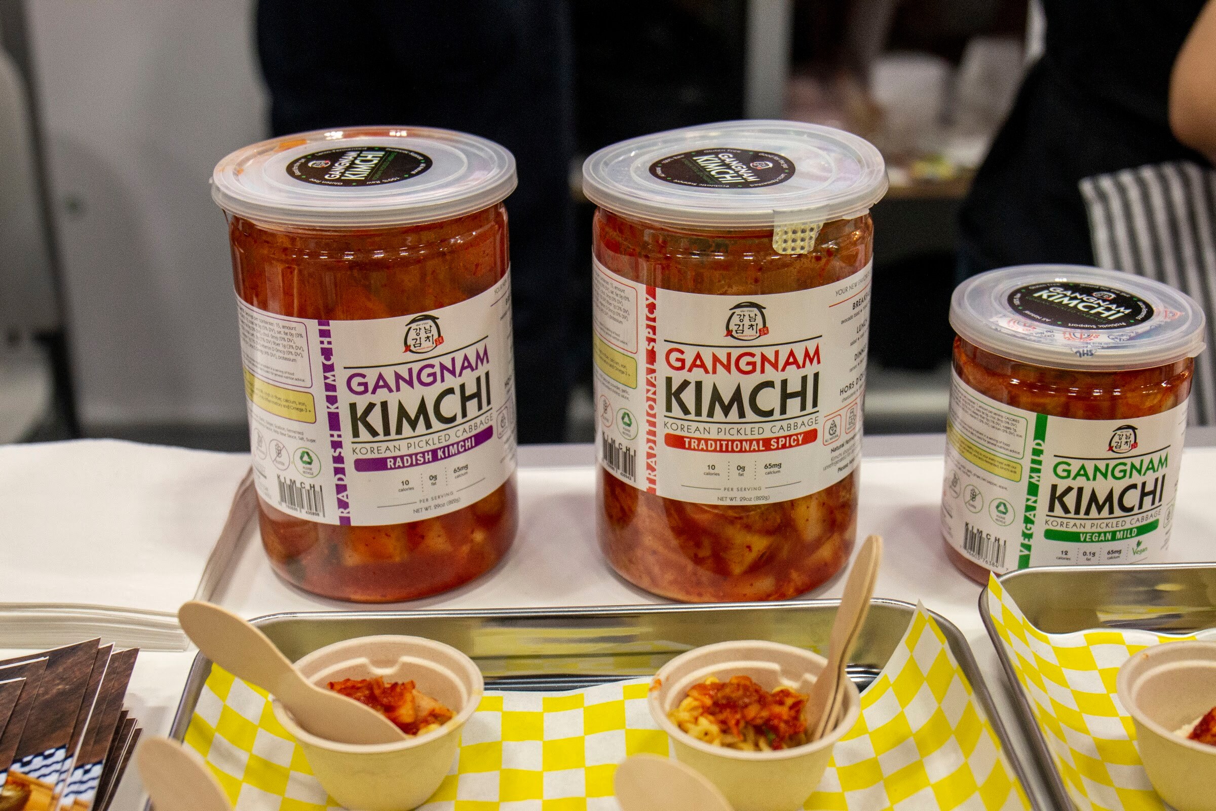 Three jars of Gangnam Kimchi in radish, traditional spicy and vegan mild with sample cups in front