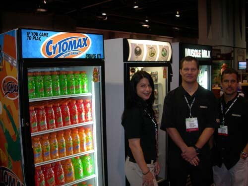 A woman and two men in black shirts stand between a Cytomax Sport Drink cooler filled with green, red and orange bottles and a Muscle Milk cooler.
