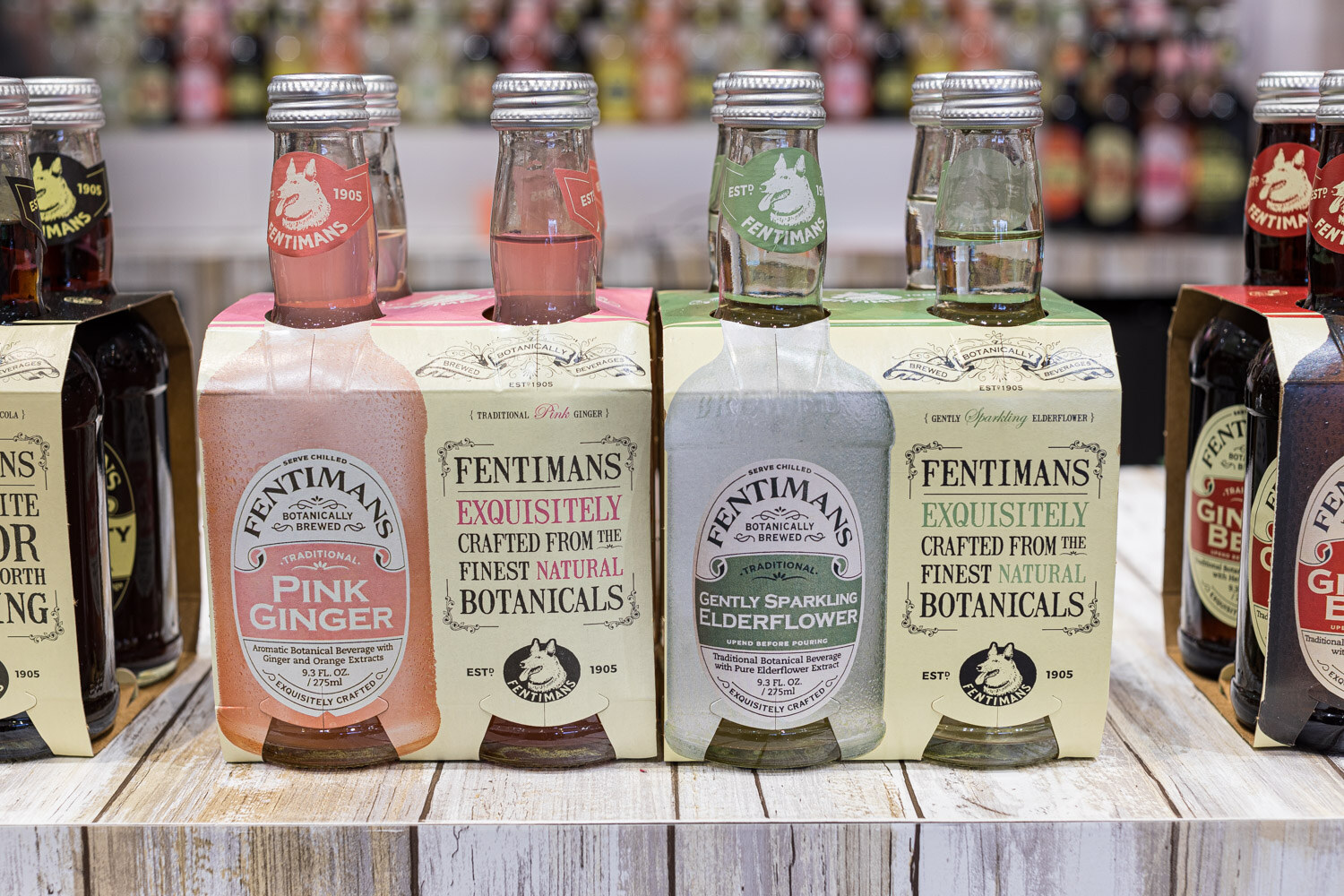 Four-packs of Fentimans Pink Ginger and Gently Sparkling Elderflower bottles on a white wooden shelf