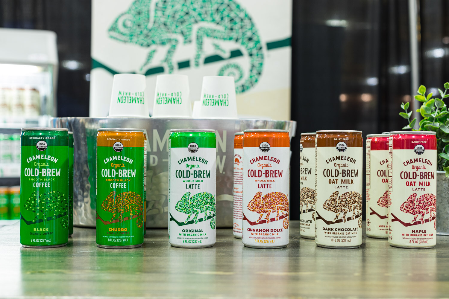 Rows of Chameleon Organic Cold-Brew cans in coffee, latte and oat milk varieties in front of a chameleon logo