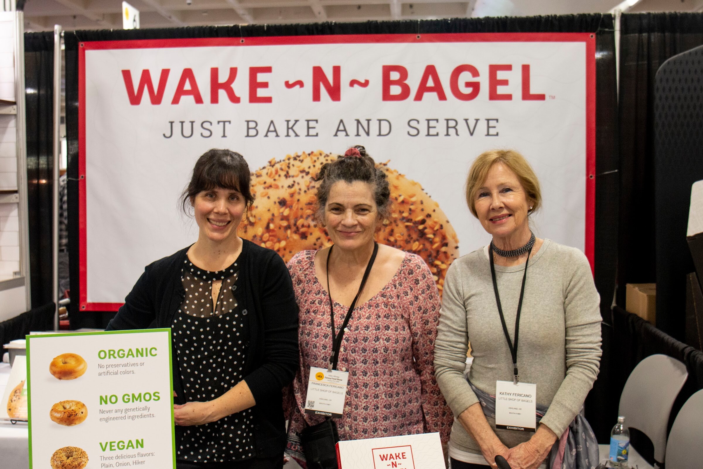 Three smiling women with badges stand in front of a Wake-N-Bagel Just Bake and Serve banner