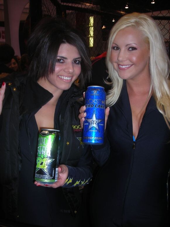 Two smiling women in black Rockstar outfits pose together; one holds a green Rockstar 21 can and the other holds a blue Rockstar Zero Carb can.
