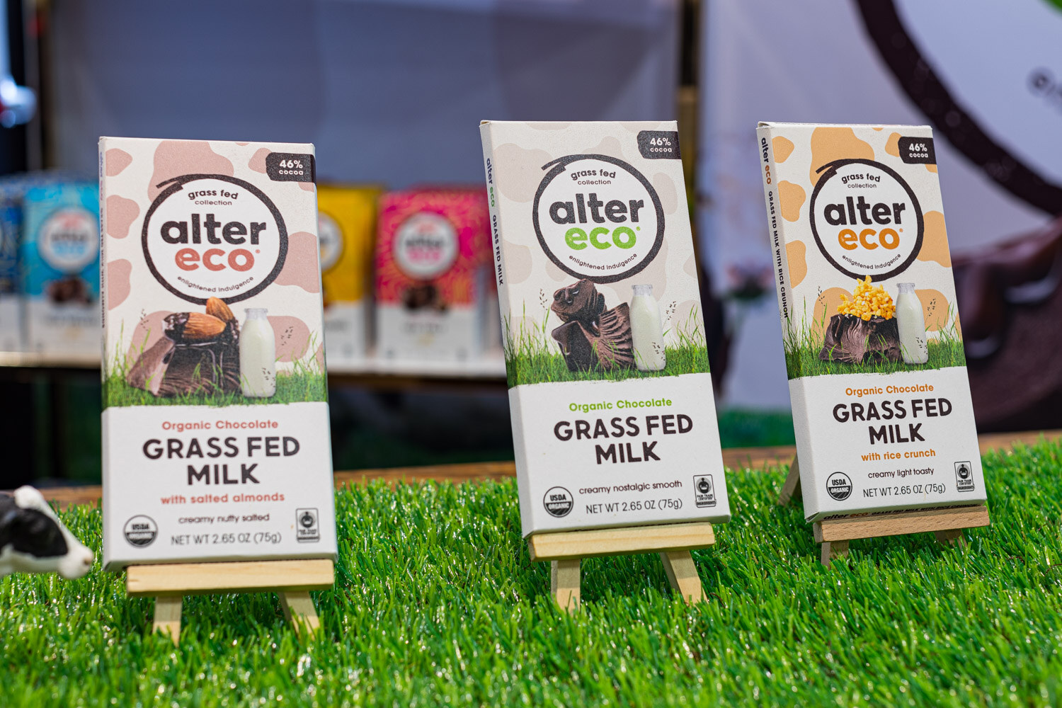 Three Alter Eco Grass Fed Milk chocolate bars on small easels on artificial grass