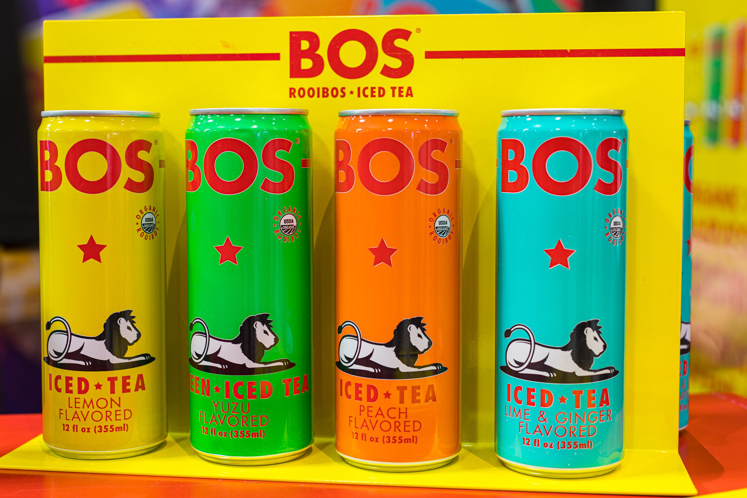 Four BOS Iced Tea cans in lemon, yuzu, peach and lime and ginger flavors on a yellow display