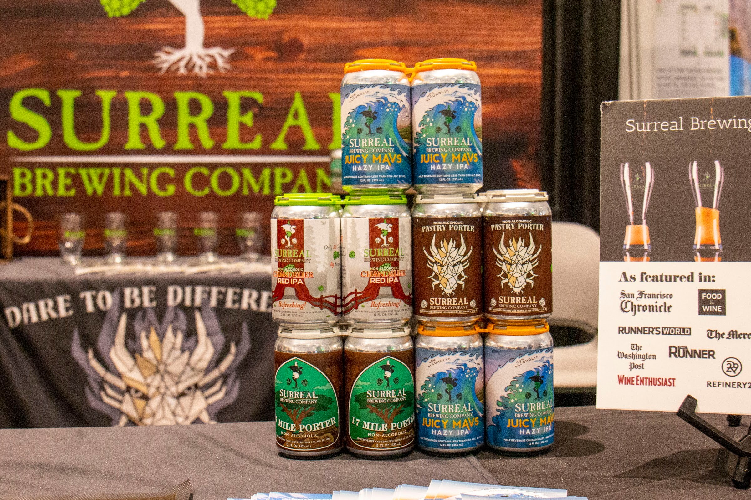 Stacked cans of Surreal Brewing Juicy Mavs hazy IPA and porters in front of a Surreal Brewing Company banner