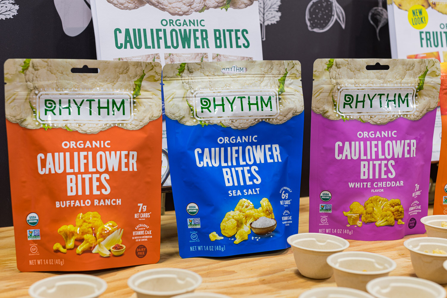 Three bags of Rhythm Organic Cauliflower Bites in orange, blue and purple with sample cups