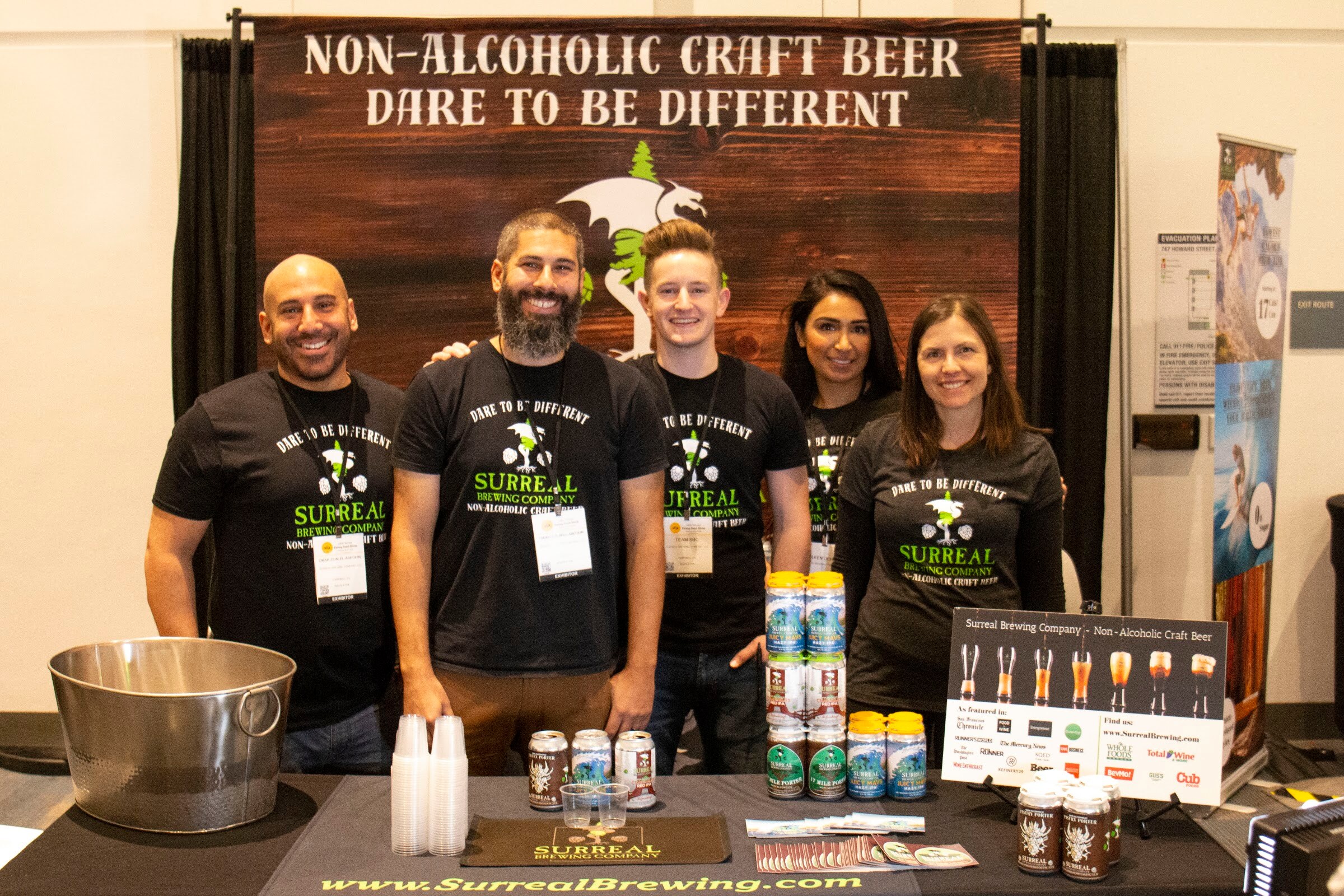 Five people in black Surreal Brewing T-shirts stand behind a table of cans under a Non-Alcoholic Craft Beer banner