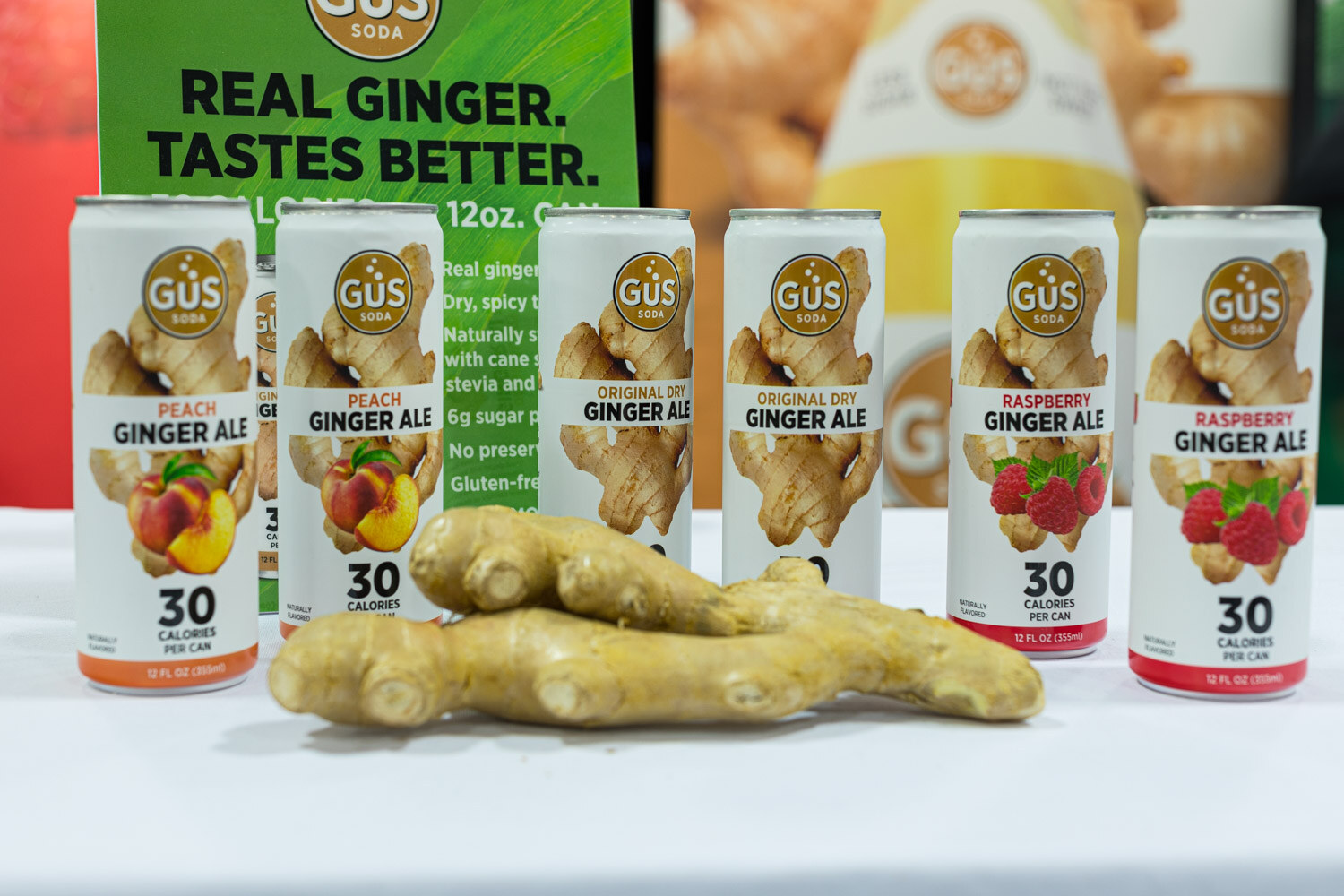 Six GUS Soda cans in peach, original dry and raspberry ginger ale beside pieces of ginger root