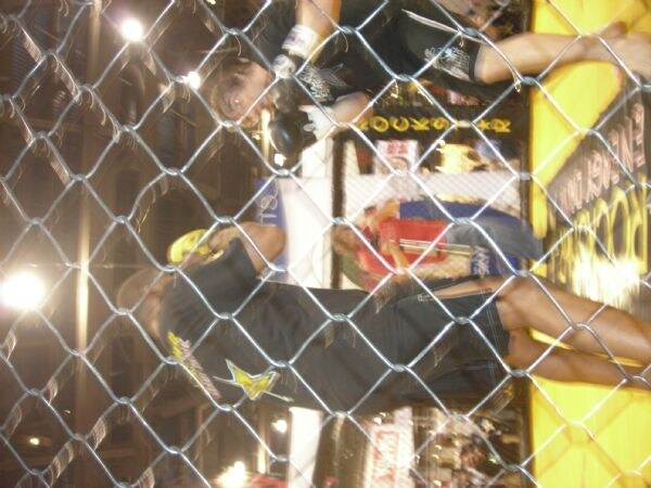 A sideways photo taken through chain-link fencing shows fighters in a ring with Rockstar Energy Drink logos on the mat and on a fighter's black shirt.