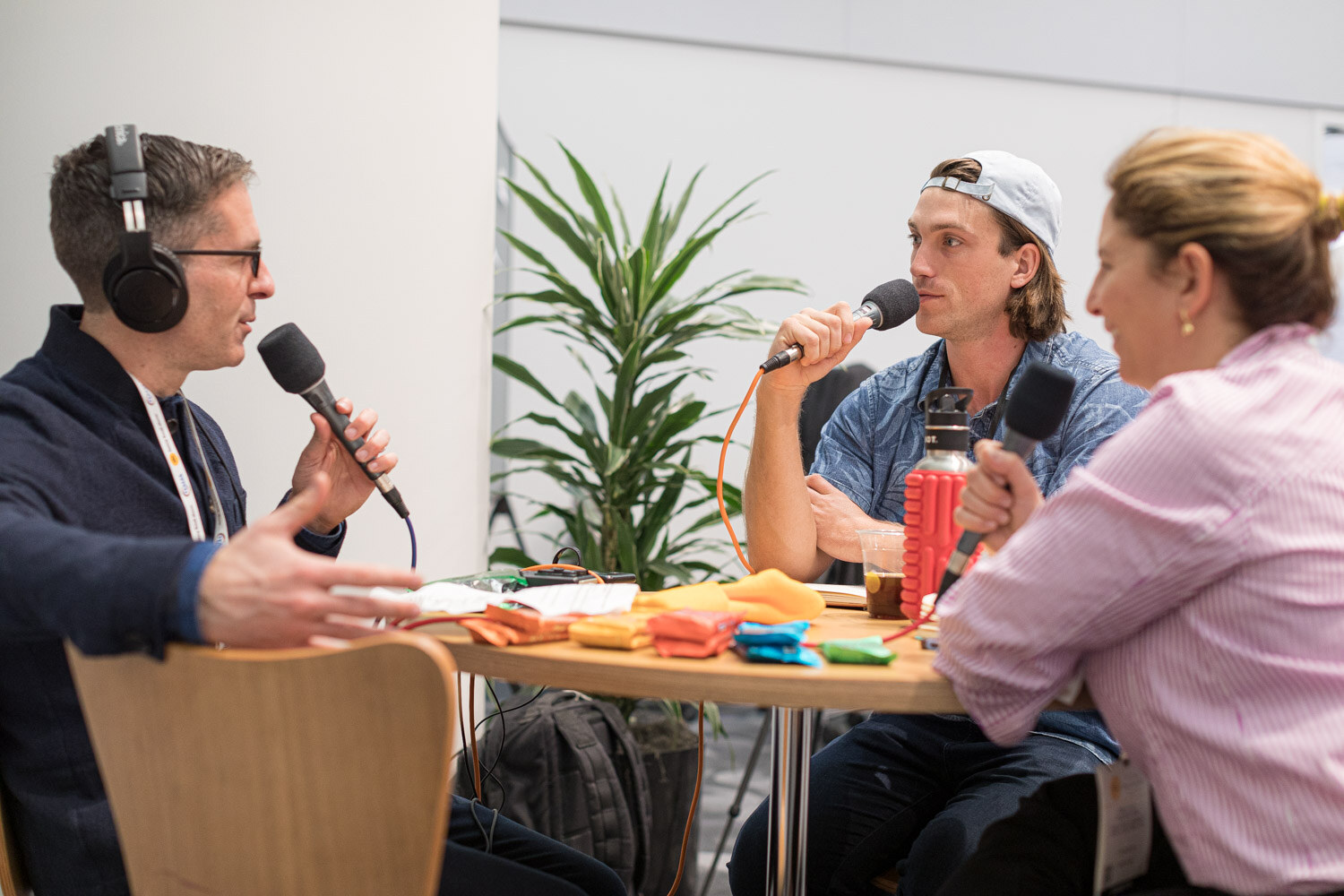 Three people holding microphones around a table, one wearing headphones, with a plant and chairs behind them