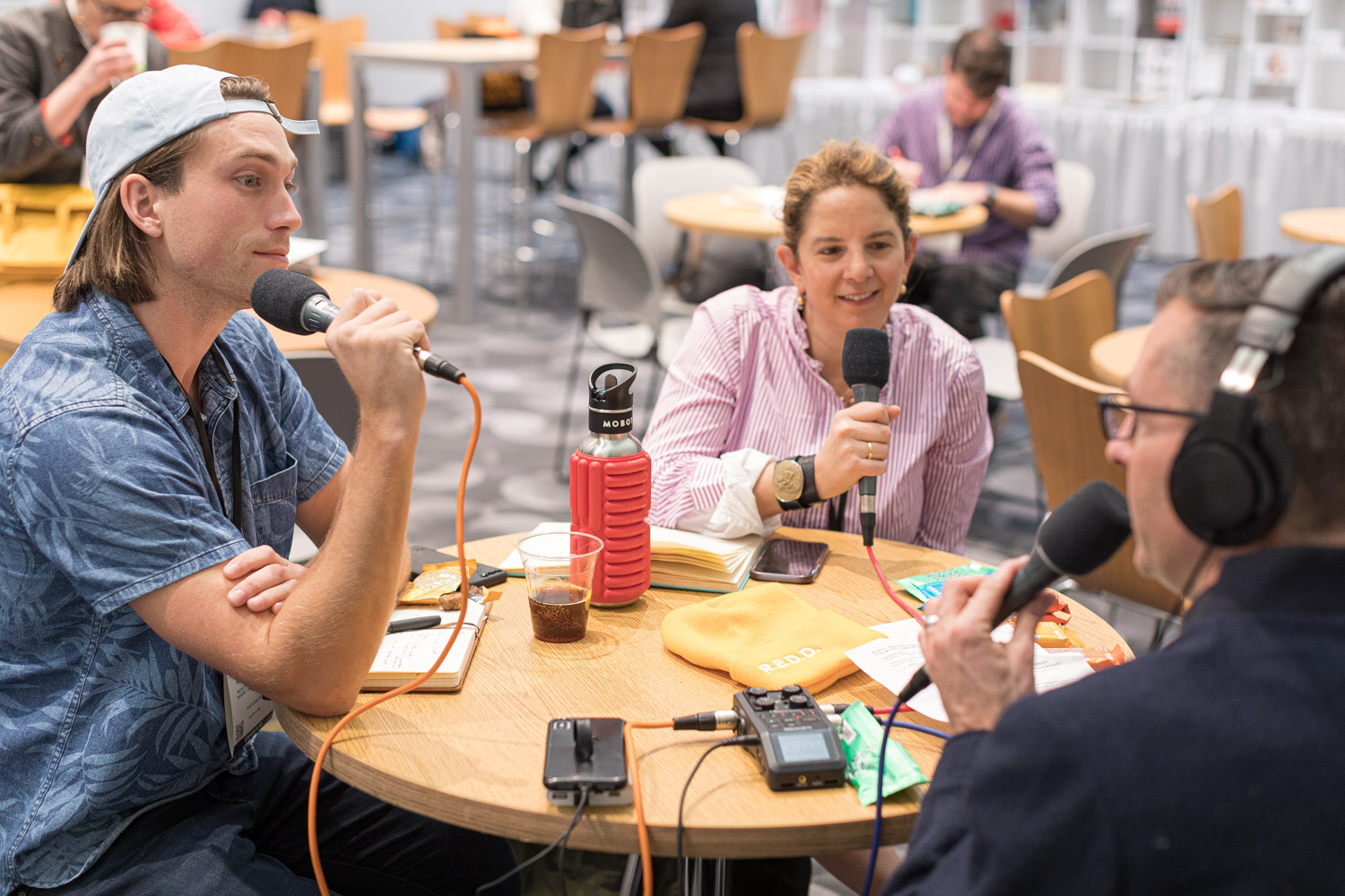 A man in a backwards cap and a woman in a striped shirt hold microphones at a small table while an interviewer in headphones sits at the right edge