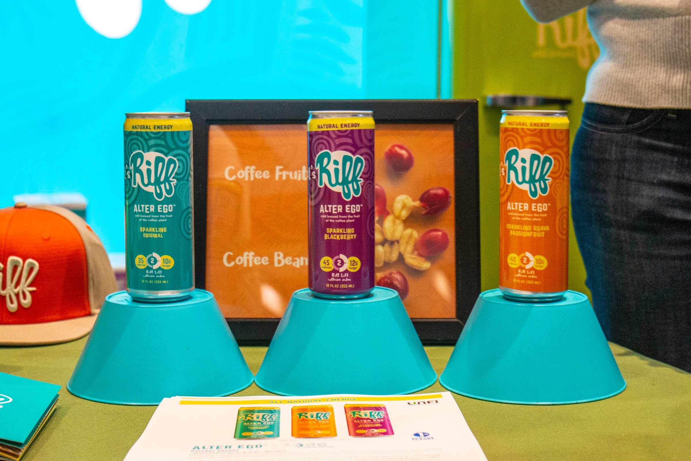 Three Riff Alter Ego cans on turquoise stands in front of a framed coffee fruit photo