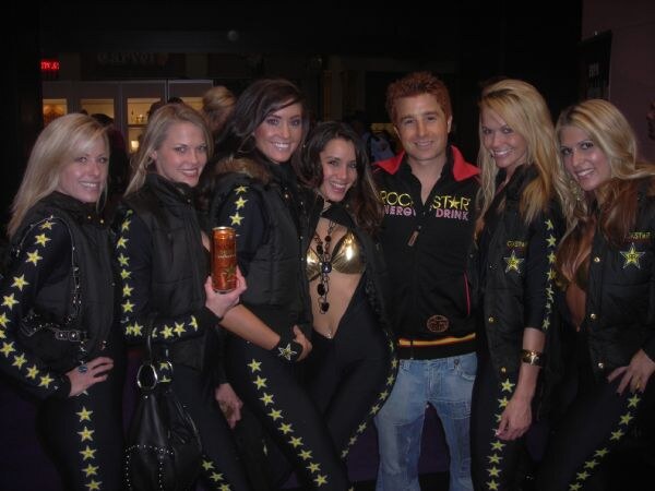 Six women in black Rockstar jackets and star-striped pants pose with a man in a black Rockstar Energy Drink jacket; one woman holds a can of Rockstar Energy Drink.