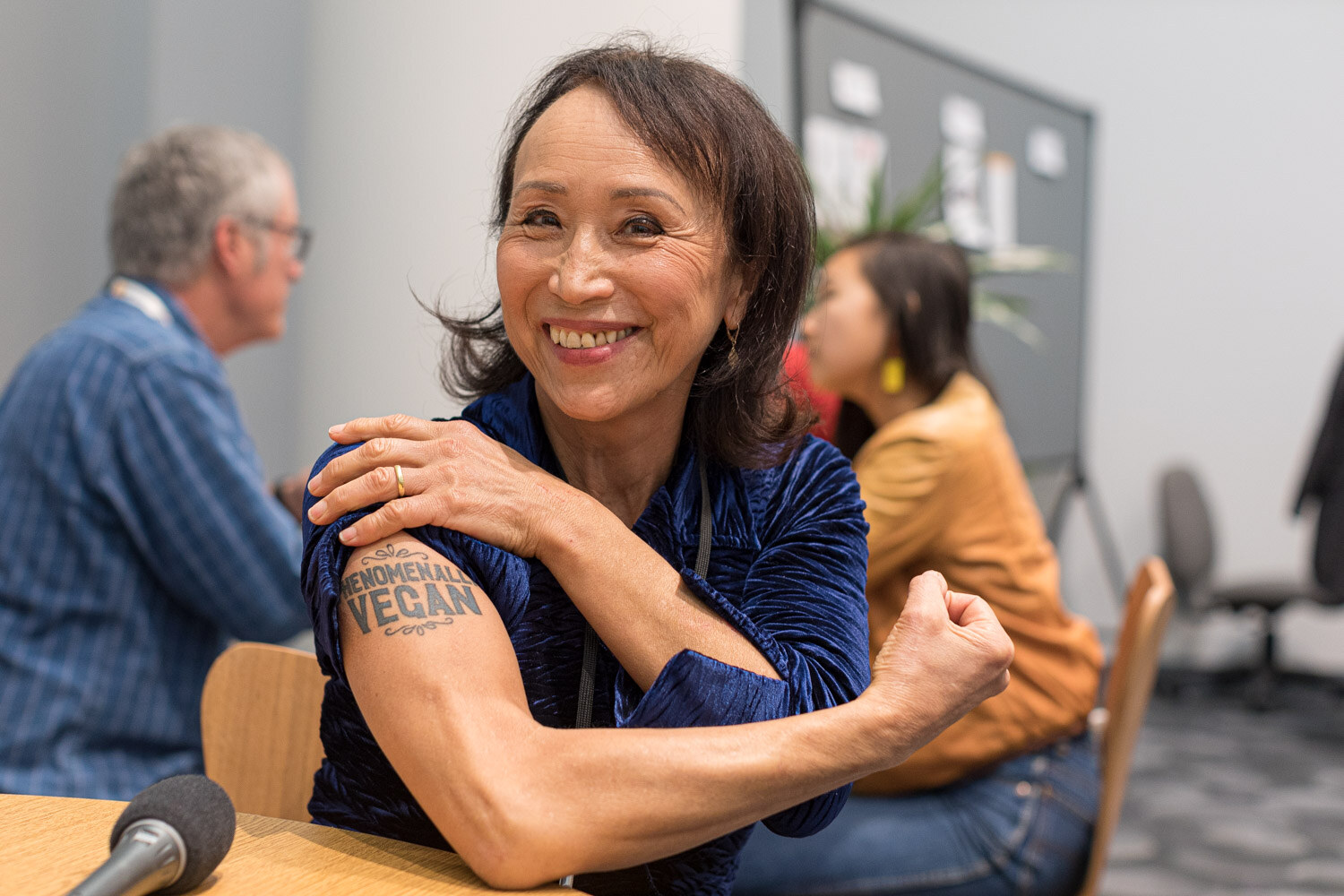 A smiling woman holds up her arm, which has a Phenomenal Vegan temporary tattoo, beside a table microphone