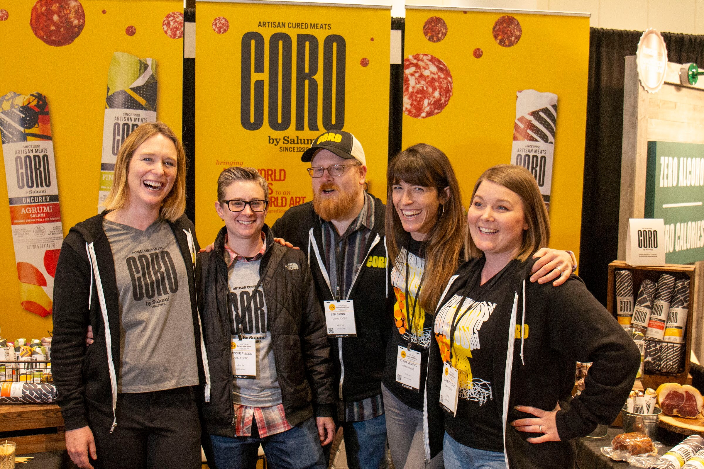 Five laughing people with badges stand in front of yellow Coro banners
