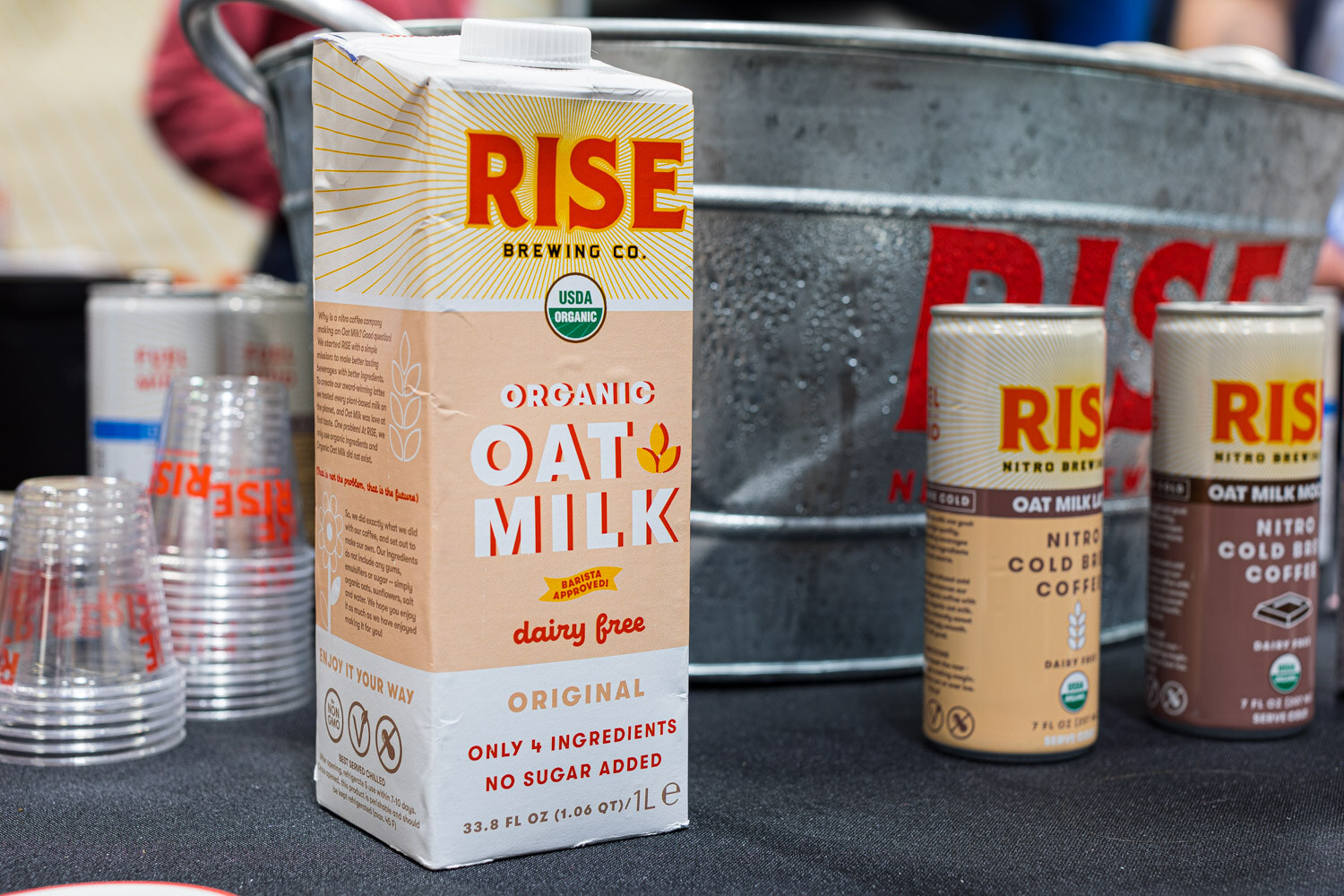 A Rise Brewing Co. oat milk carton and two Rise nitro cold brew cans in front of a large Rise tub