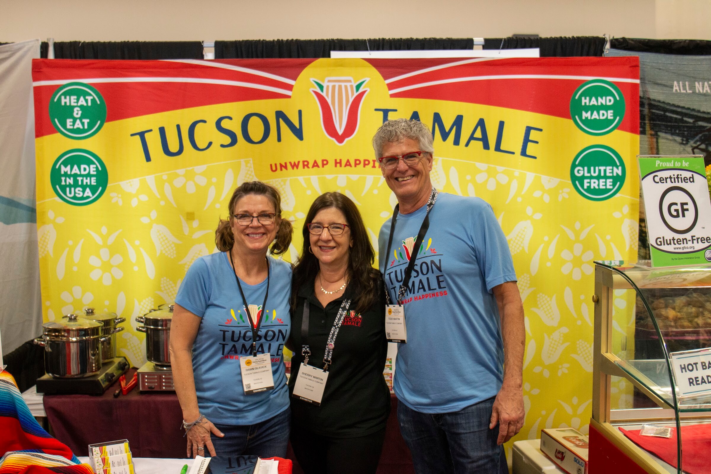 Three people in Tucson Tamale shirts stand in front of a yellow Tucson Tamale backdrop with cooking pots beside them