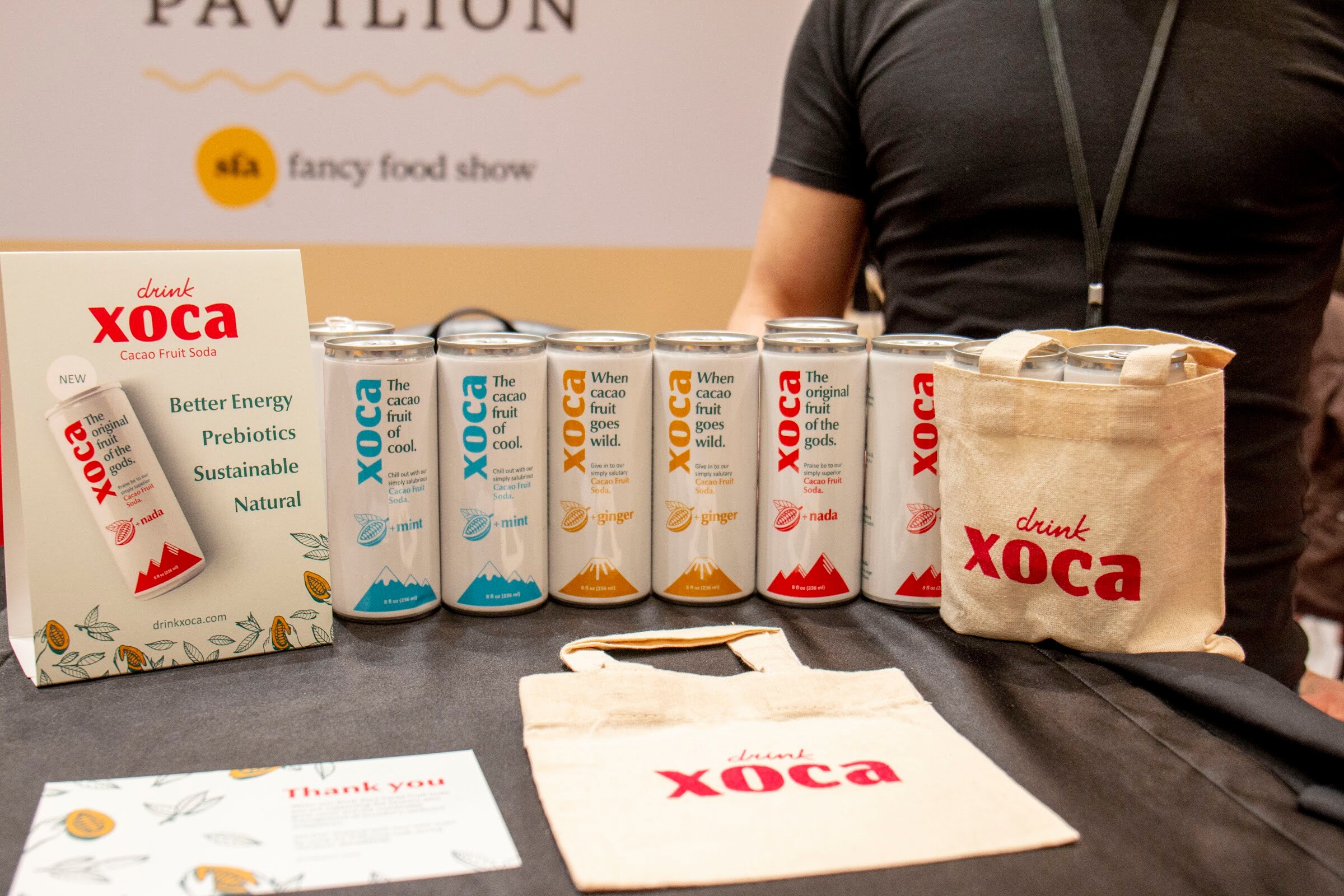 Xoca cans in several flavors with tote bags and a Drink Xoca sign under a Fancy Food Show pavilion sign