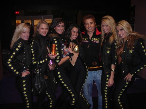Six women in black star-patterned Rockstar outfits stand beside a man in a black Rockstar Energy Drink jacket and jeans; a couple of them hold cans.