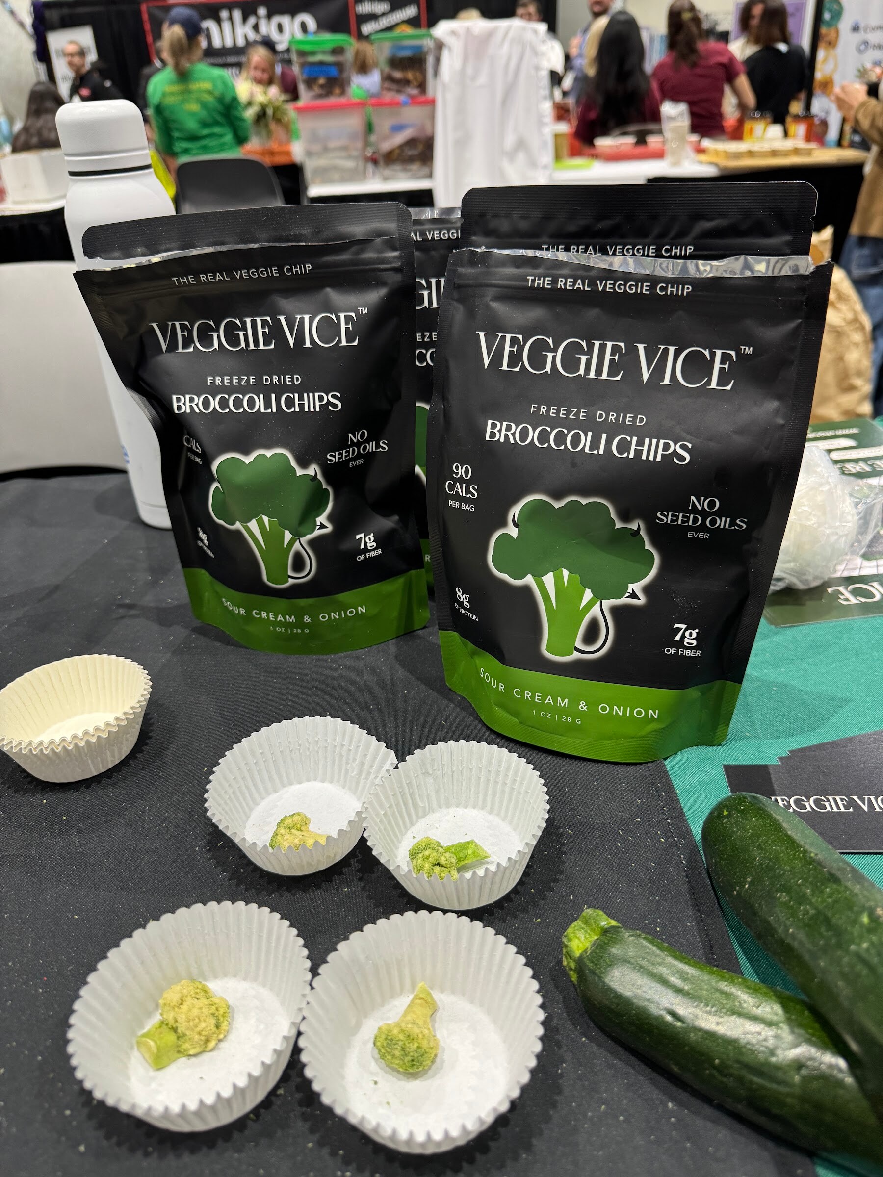 Bags of Veggie Vice freeze-dried broccoli chips with sample cups and fresh zucchini on a dark table.