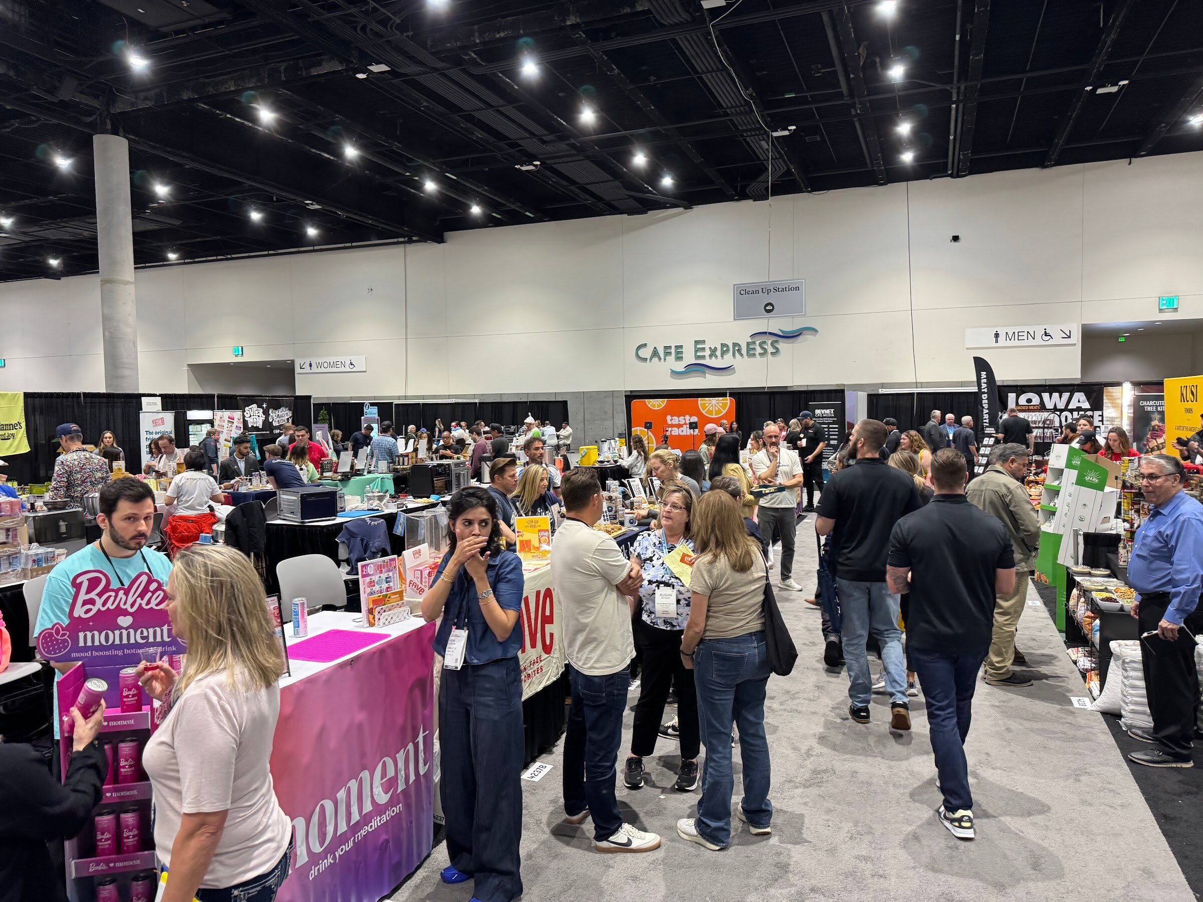 Busy trade show aisle with crowds of attendees between booths under convention center lights.
