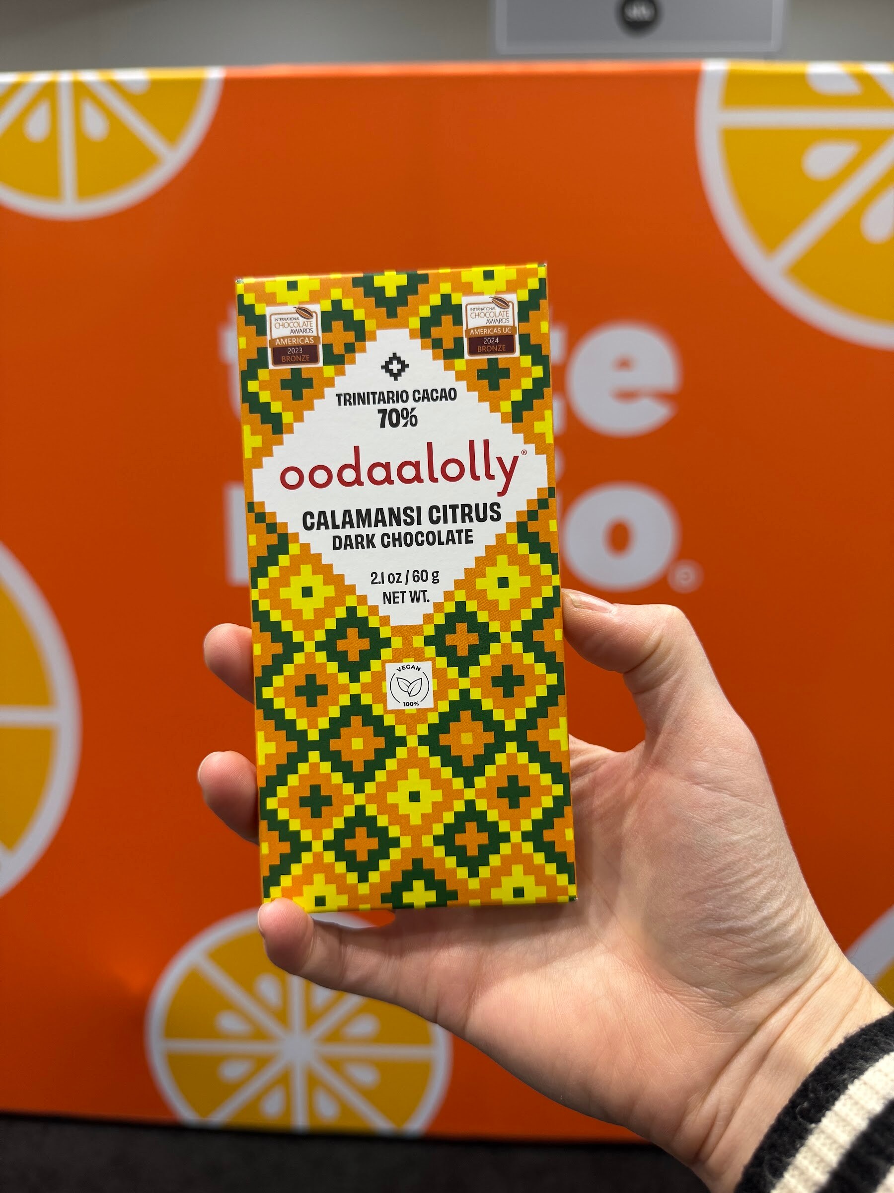 Hand holds a patterned oodaalolly calamansi citrus dark chocolate bar against an orange backdrop.