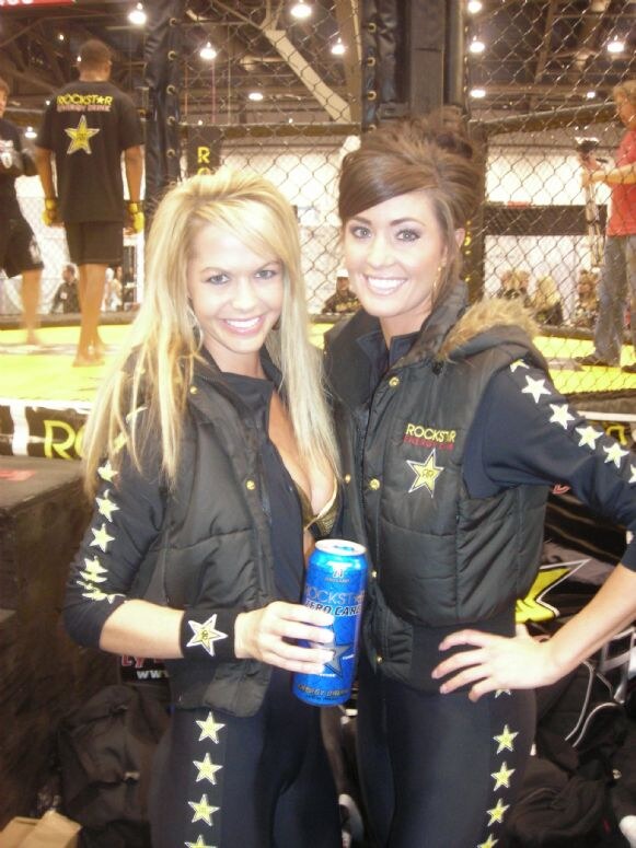 Two smiling women in black Rockstar vests and star-striped pants stand before a caged fighting ring; one holds a blue can of Rockstar Energy Drink Zero Carb.